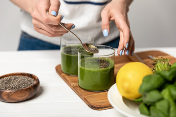 cropped view of woman holding spoon with chia seeds near glasses of green fresh smoothie on grey