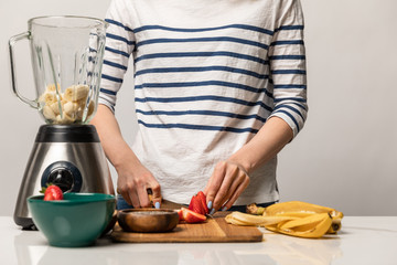 cropped view of woman cutting sweet strawberries near bananas and blender on white