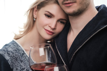 A couple, a man and a woman in bathrobes drink red wine from large glass goblets in the apartment. Kiss and hug.