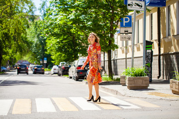 Young beautiful blonde woman in a red flower dress crosses the road at a crosswalk