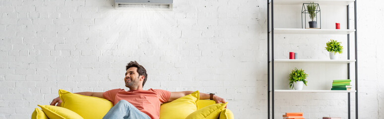 panoramic shot of handsome man sitting on yellow sofa under air conditioner at home