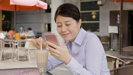 pretty asian woman employee holding modern beverage bubble milk tea sitting in outdoor coffee shop on sunny day. happy smiling young female manager typing text message on smart phone break time