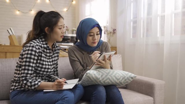 two young female multi student study together making notes working on notebook. asian chinese classmate discussing talking with arabian girl prepare for exam test writing sitting on sofa in dormitory