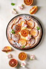 Homemade Cheesecake with sicilian blood oranges, decorated by edible flowers, mint leaves and sugar powder served in plate with cutted oranges above over white marble background. Flat lay, space