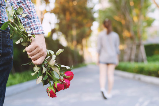 Young Man Standing With A Red Rose On Hand Sadness Love In Ending Break Up Of Relationship Blurred Woman Back Side Walking Away Parting  In Public Park Outdoor.  Broken Heart In Valentine Day Concept.