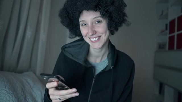 Afro haired white girl e mailed, chat with her smart phone and smiling to camera in her room. Curly haired happy woman