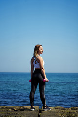 Vertical photo of a girl who does sports exercises on the beach of the ocean or the sea. The girl is standing and holding dumbbells in her hands.