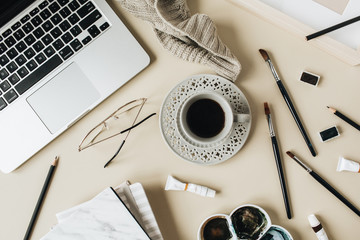 Watercolor artist home office desk workspace on beige background. Laptop, coffee cup, paint brushes, plaid, painting, watercolor. Flat lay, top view blog, social media, website template.