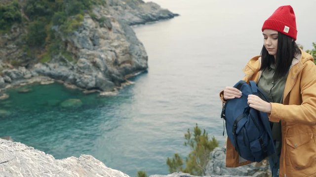 Tourist girl enters the frame with a backpack and sits on the edge of the cliff. Enjoys a beautiful view on a cloudy autumn day. 4K