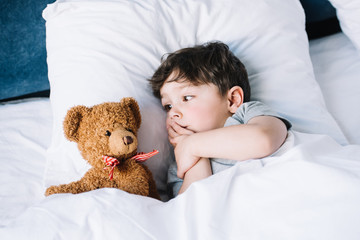 cute kid lying on white pillow in bed and looking at teddy bear