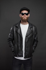 Man wearing leather biker jacket or asymmetric zip jacket with black cap, jeans and sunglasses. Handsome hipster man over gray background