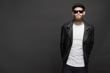 Man wearing leather biker jacket or asymmetric zip jacket with black cap, jeans and sunglasses. Handsome hipster man over gray background