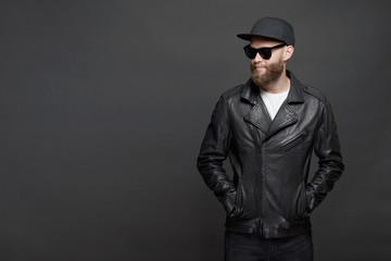 Obraz premium Man wearing leather biker jacket or asymmetric zip jacket with black cap, jeans and sunglasses. Handsome hipster man over gray background