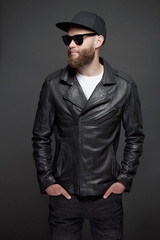 Obraz premium Man wearing leather biker jacket or asymmetric zip jacket with black cap, jeans and sunglasses. Handsome hipster man over gray background