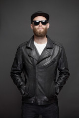 Obraz premium Man wearing leather biker jacket or asymmetric zip jacket with black cap, jeans and sunglasses. Handsome hipster man over gray background