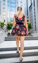 Obraz premium Toned rear view image of sexy young woman in short dress and ballet flats walking up the stone stairs next to high modern building and skyscrapers