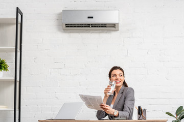 cheerful businesswoman drinking water while sitting at workplace under air conditioner