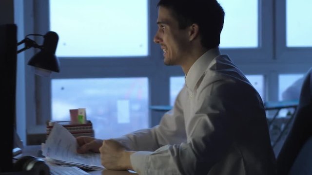 Angry businessman in formalwear working on computer in office at night, then throws papers. 3840x2160