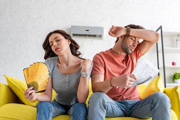 Fotobehang pretty woman with hand fan and handsome man with newspaper suffering from heat at home © LIGHTFIELD STUDIOS