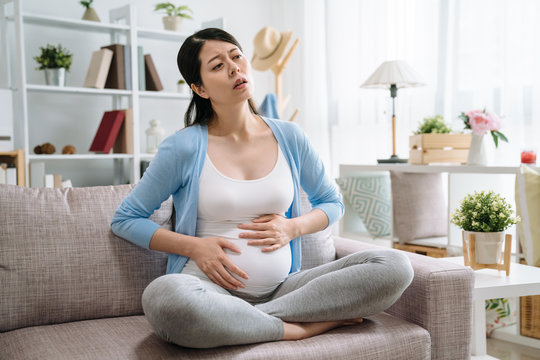 Young pregnant woman suffering from abdominal pain at home. beautiful asian girl future parent belly hurting painful sit on couch sofa at home in cozy bright modern apartment. chinese female illness
