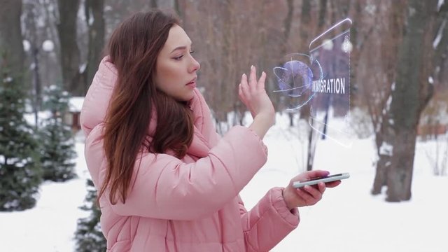 Beautiful young woman in a winter park interacts with HUD hologram with text Immigration. Red-haired girl in warm pink clothes uses the technology of the future mobile screen