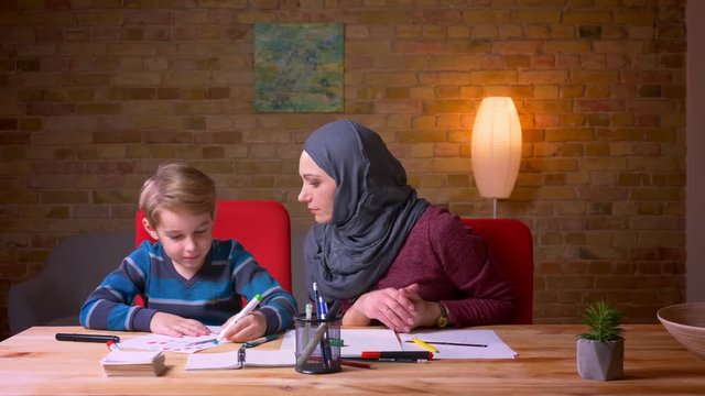 Muslim mother in hijab helping her son draw the picture with markers at the table in cozy home atmosphere.