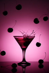 Pink cherry coctail in martini glass with a splash and falling cherries on the background