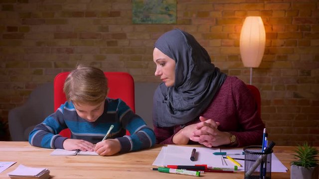 Muslim mother in hijab helps her son do the homework at the table.