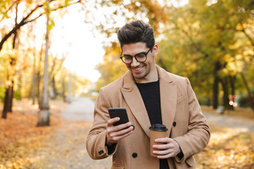 Image of handsome casual man wearing coat using cellphone and smiling n autumn park