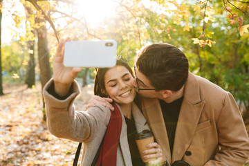Portrait of smiling couple drinking takeaway coffee nd taking selfie photo on cellphone while...