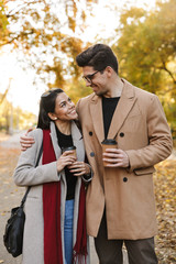 Fototapeta premium Portrait of young couple drinking takeaway coffee and looking at each other while walking in autumn park