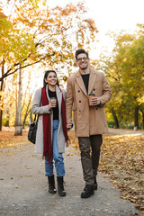 Portrait of casual couple drinking takeaway coffee from paper cups while walking in autumn park