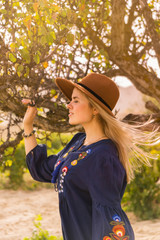 Beautiful blond hair young woman wearing blue skirt and brown hat near  lonely tree  in grapeyard - plantation of grapevines. Spring dawn solar bright warm effect.