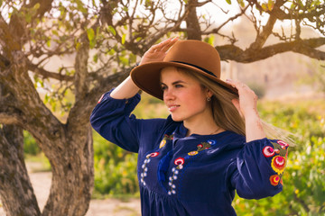 Beautiful blond hair young woman wearing blue skirt and brown hat near  lonely tree  in grapeyard - plantation of grapevines. Spring dawn solar bright warm effect.