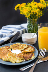 zucchini pancakes with sour cream on a plate, a glass of orange juice ,a bouquet of yellow flowers, dandelions, towel, fork ,spoon