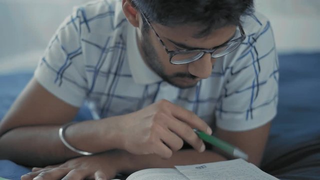 Close-Up, Indian Student in Glasses Thoughtfully Reads Entries in the Lay Lie on the Bed