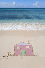 Fototapeta premium A house drawn in the sand on a beautiful sandy beach with waves lapping the shore. Symbol of moving house, moving overseas, new life.