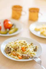 Macaroni with slices of meat and carrots. Marinated tomatoes and cucumbers. Cheese sliced in a plate. Two mugs on the table