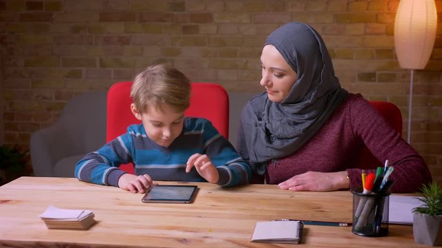 Small boy watching into tablet and his muslim mother in hijab sitting near at home.