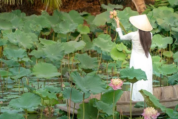 Fototapete Beautiful asia women wearing white white traditional Vietnam dress (Ao Wai) and Vietnam farmer's hat and standing on wooden boat in flower lotus lake. Her hands holding lotus flowers. © Prot