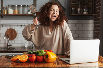 Image of joyful caucasian woman using laptop while cooking in kitchen interior at home