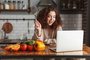 Image of smiling caucasian woman using laptop while cooking in kitchen interior at home