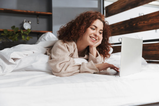 Image of cute young woman laptop while lying in bed on white linen at home