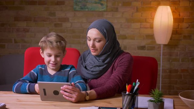 Portrait of small boy and his muslim mother in hijab watching cartoons on tablet together and discussing them.