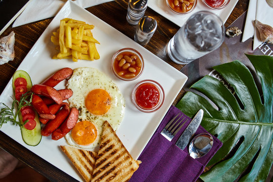 Food photography composition aesthetic. Breakfast meal on a white plate - fried eggs, chips, toasts, fried sausages and vegetables with beans and ketchup. Detailed background