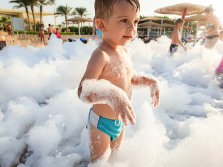 Funny portrait of little toddler boy covered in soap foam playing on the sea beach during disco...