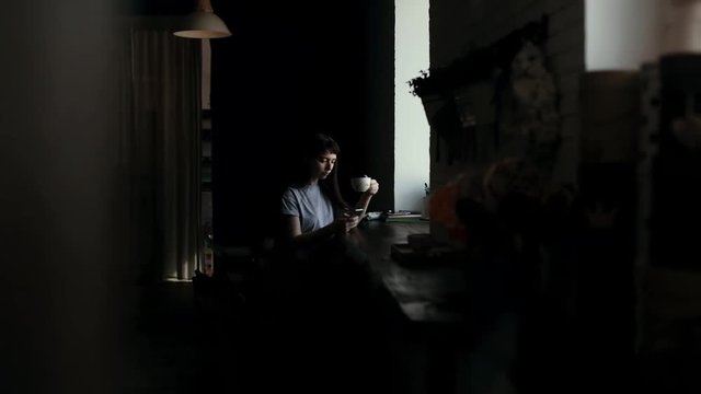 Girl sits by window, drinks coffee from a cup and typing message on phone in slow motion. Camera moves horizontally. Someone is spying on young woman who drinks sitting at table in cafe and texting