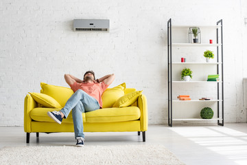 young man sitting on yellow sofa under air conditioner in spacious apartment