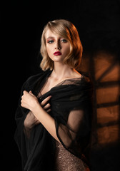 Cute face blonde girl with vintage style hairstyle, wearing a golden sparkling dress and black veil on her shoulders stands on the background of a wall with window shadow. Healthy skin