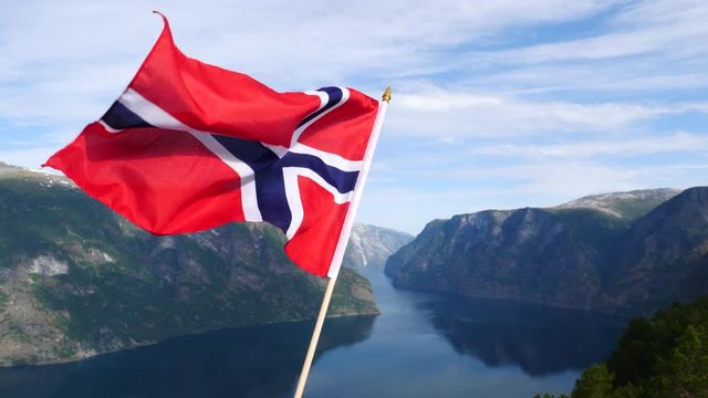 Norwegian flag waving in slow motion against fjord Aurlandsfjord landscape. View from Stegastein viewpoint. National tourist route Aurlandsfjellet